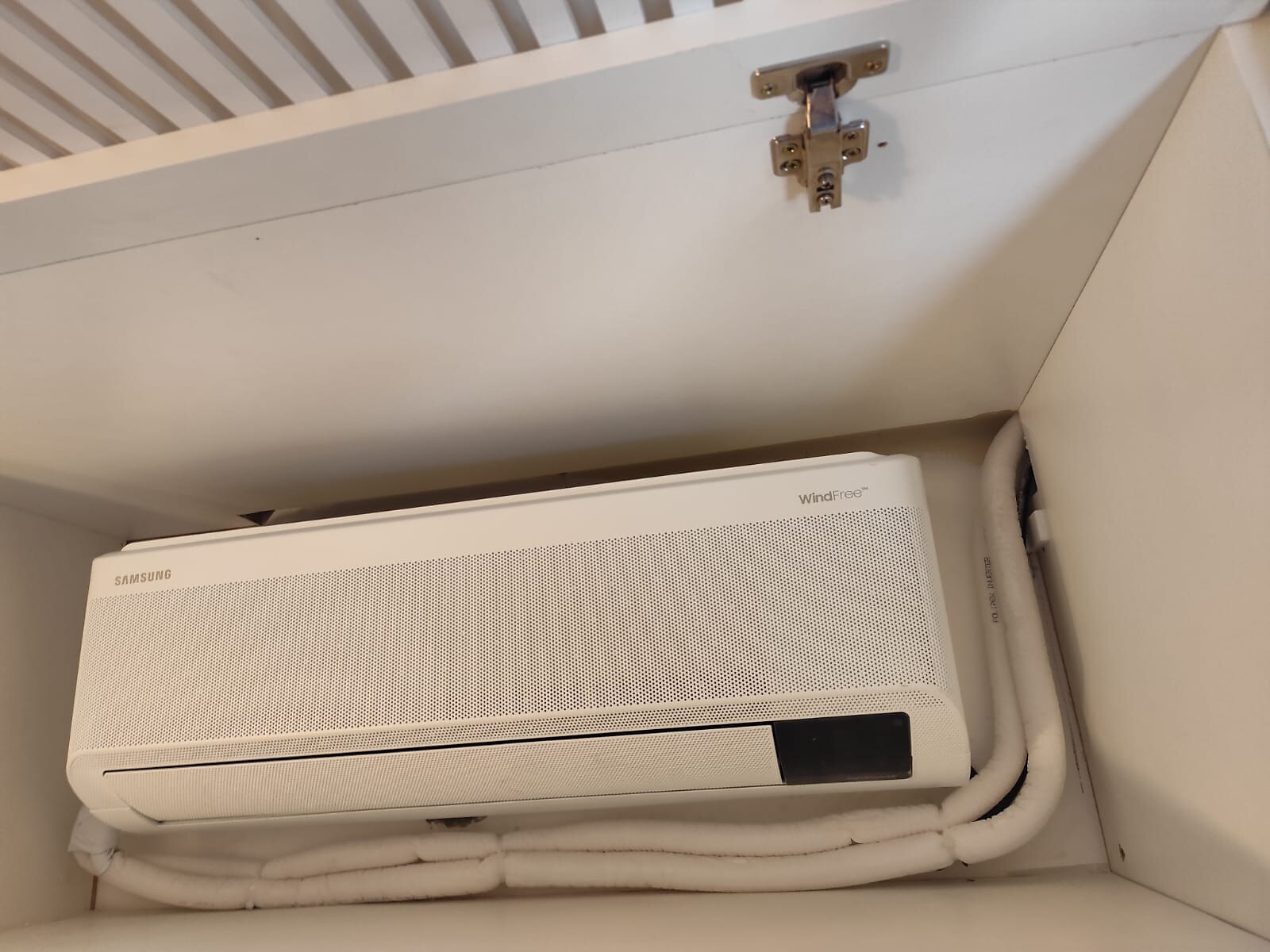 Evaporadora de ar-condicionado embutida em nicho de marcenaria com porta ripada aberta, mostrando a tubulação com isolamento térmico